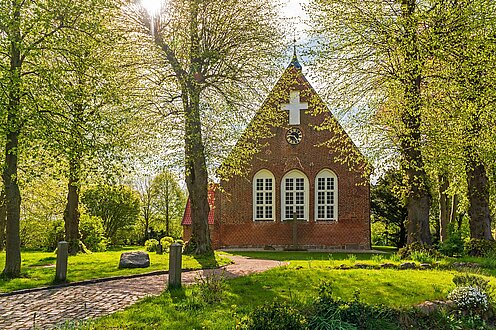 Eine kleine Kirche umgeben von grünen blühenden Bäumen, Die Sonne scheint durch die Bäume.
