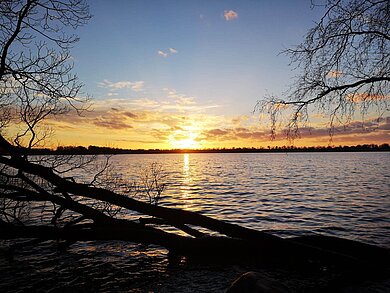 Die Sonne geht orange leuchtend über dem Wasser eines Sees unter.