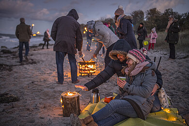 Eine große Gruppe erlebt die Fackelwanderung am Hohwachter Strand. Es ist dunkel, alle haben sich warm angezogen. Überall stehen Fackeln und vorne im Bild ein Feuerkorb, wo sich ein Paar aufwärmt.
