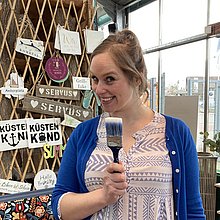 Eine Frau lacht in die Kamera, in der Hand ein Pinsel.