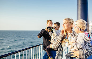 Eine Familie mit zwei Kindern ist auf dem Außendeck an der Reling zu sehen. Sie schauen auf die Ostsee. Der Junge der auf dem Arm des Vater ist, schaut durch ein Fernglas.
