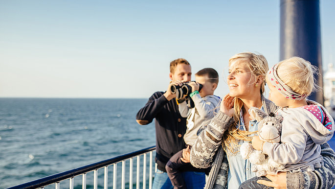 Eine Familie mit zwei Kindern ist auf dem Außendeck an der Reling zu sehen. Sie schauen auf die Ostsee. Der Junge der auf dem Arm des Vater ist, schaut durch ein Fernglas.