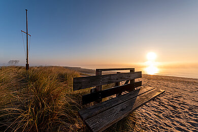 Eine leere Bank am Strand, dahinter das Meer. Die Sonne steht blutrot am Horizont. Der Himmel ist klar. 