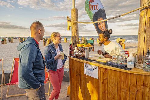 Eine Cocktailbar direkt am Strand. Auf dem Bild sind ein Mann und eine Frau, die gerade etwas bestellen. Hinter ihnen das Meer. Es ist Abendlicht. 