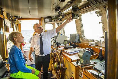 Ein Junge sitzt in einem Schiff in der Fahrerkabine und der Kapitän zeigt ihm die Ausstattung.
