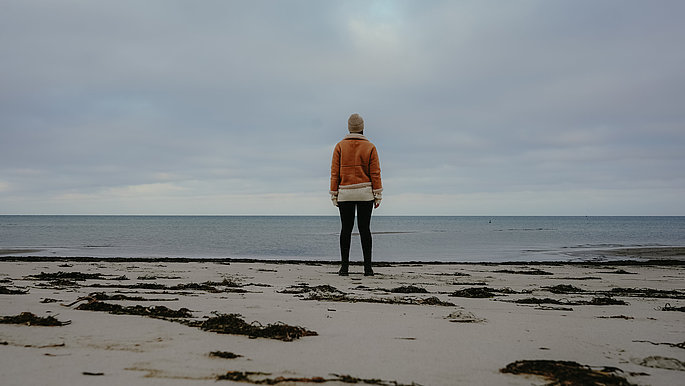 Ein einsamer Winterstrand, zu sehen eine Frau von hinten, die auf das Meer blickt