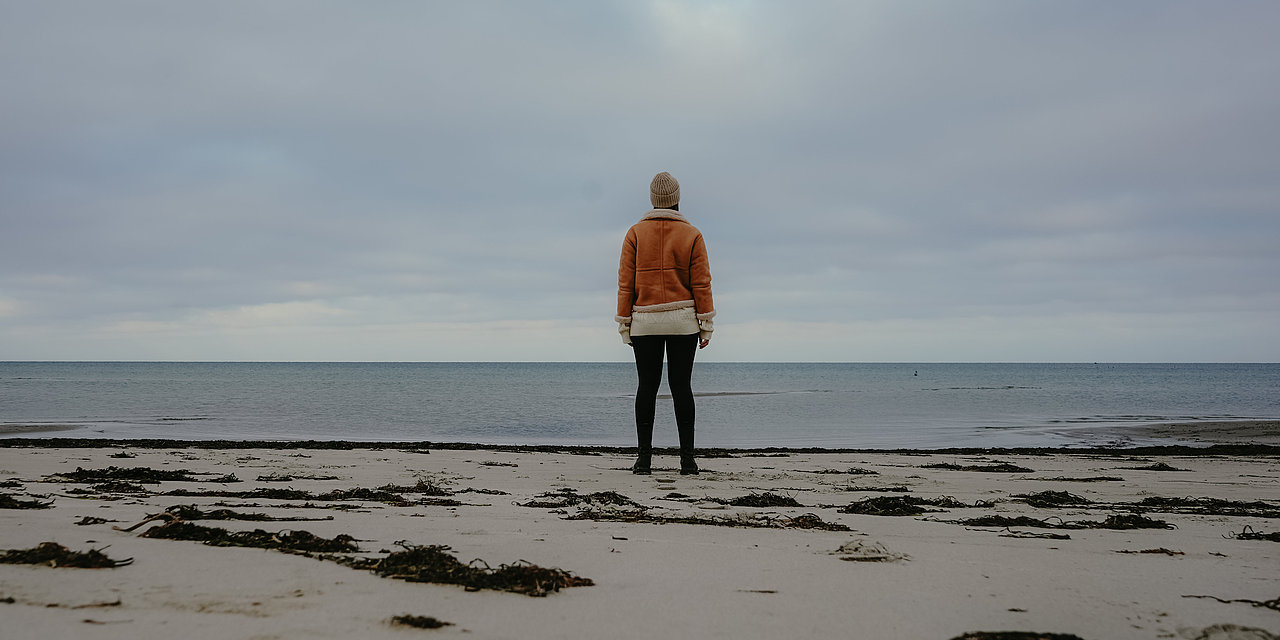 Ein einsamer Winterstrand, zu sehen eine Frau von hinten, die auf das Meer blickt