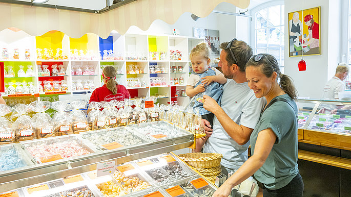 Familie mit Kleinkind besucht die Bonbonkocherei. Der Vater trägt das Kind auf dem Arm. Zusammen bestaunen sie die Auswahl an Bonbons.
