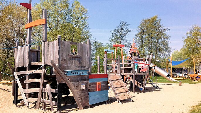 Piratenlager Hohwacht Spielplatz. zu sehen ist ein hölzernes Piratenschiff zum Klettern auf Sand. Im Hintergrund ein paar Bäume.