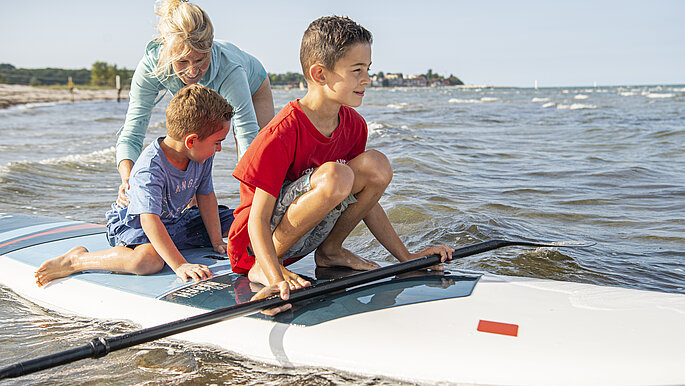 Eine Frau schiebt zwei Kinder, die auf einem SUP sitzen, über das Wasser.