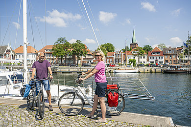 Zwei Personen mit jeweils einem Fahrrad stehen am Hafen von Neustadt in Holstein. Hinter ihnen sieht man das Hafenbecken mit weißen Segelbooten.