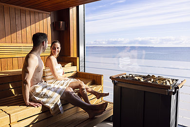 Paar sitzt in der Sauna des Bayside Hotels und schaut auf die Ostsee. Vor dem Fenster seht der Saunaofen