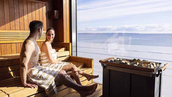 Paar sitzt in der Sauna des Bayside Hotels und schaut auf die Ostsee. Vor dem Fenster seht der Saunaofen
