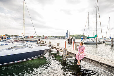 Frau sitzt auf Steg am Wasser. Neben ihr viele Segelboote im Hafen. 