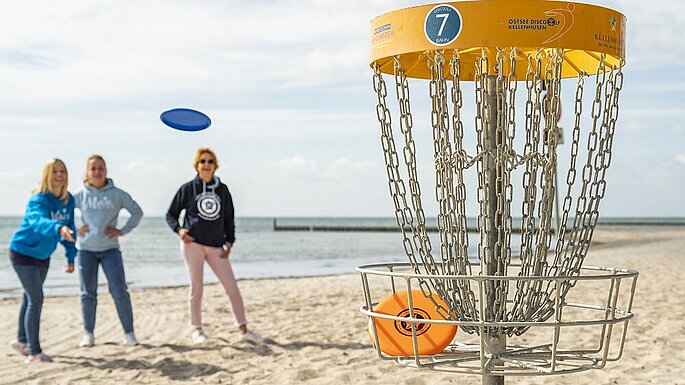 Eine Wurfscheibe fliegt durch die Luft. direkt am Strand steht das Netz aus Ketten, in das die Scheibe fliegen soll. Im Hintergrund das Meer.