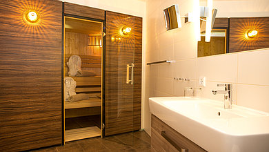 Zu sehen ist ein Wellness-Zimmer. Im Hintergrund ist eine Sauna zu sehen und im Vordergrund auf der rechten Seite ein weißes Waschbecken.