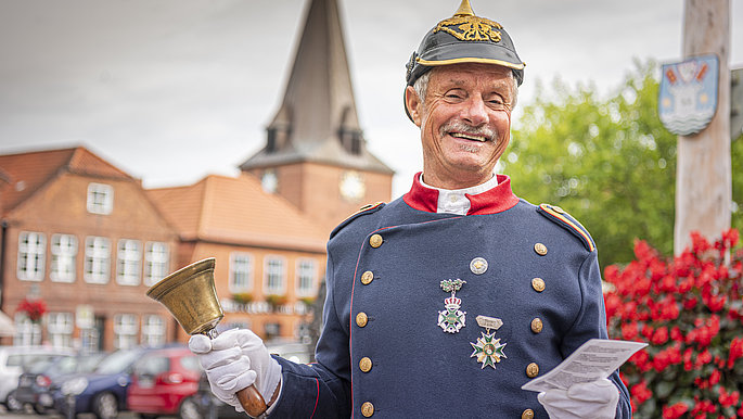 Eine Mann, verkleidet als eine Figur aus einem vorherigen Zeitalter, gibt eine Stadtführung in Lütjenburg. Er steht auf dem Marktplatz mit der Kirche im Hintergrund.