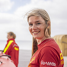 Frau in roter DLRG-Kleidung am Strand, im hintergrund ein Team mit Schlauchboot