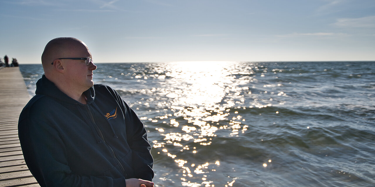 Michael Plambeck ist Saunameister im FehMare. Er steht auf der an dem Steg der ein paar Meter auf die Ostsee führt und schaut glücklich und zufrieden bei Sonnenschein auf die Ostsee 