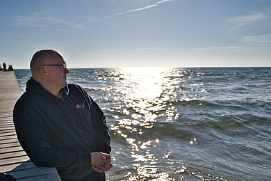 Michael Plambeck ist Saunameister im FehMare. Er steht auf der an dem Steg der ein paar Meter auf die Ostsee führt und schaut glücklich und zufrieden bei Sonnenschein auf die Ostsee 