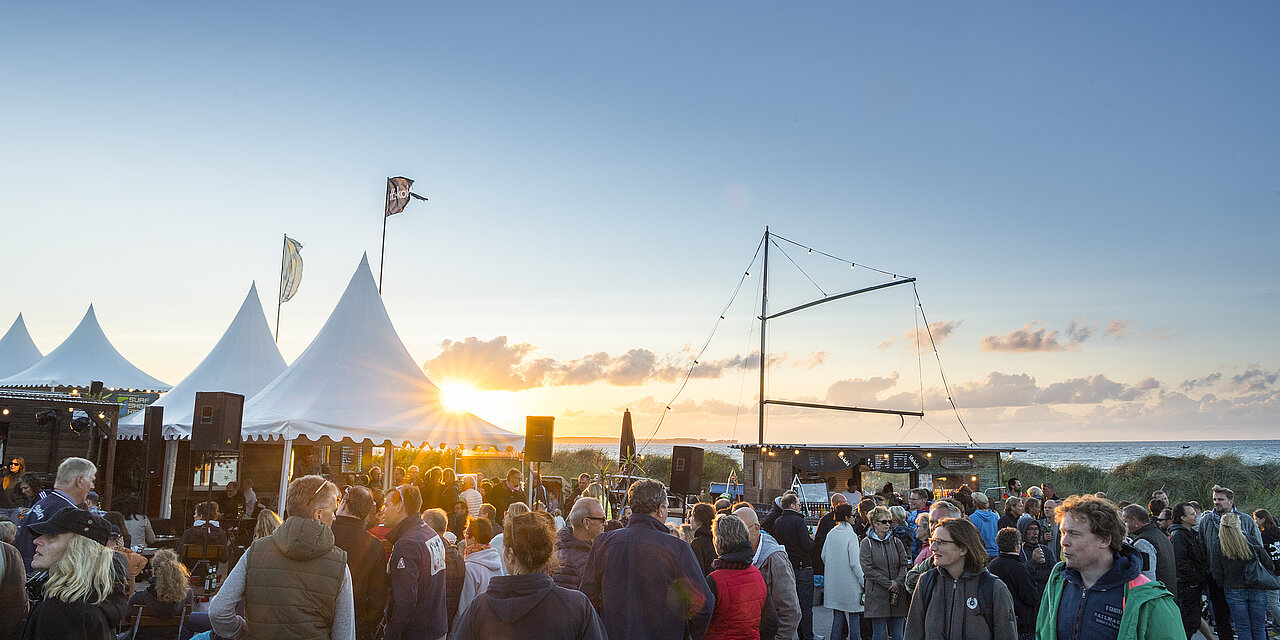 Viele Menschen sind auf einer Musikveranstaltung beim Tatort Hawaii in Stein. Hinter den weißen Zelten ist der Sonnenuntergang an der Ostsee zu sehen.