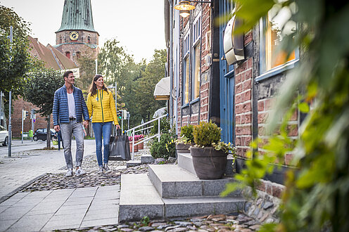 Eine Altstadt. Rechts im Bild ein Hauseingang, im Hintergrund eine Backsteinkirche, zwei Menschen gehen spazieren. 