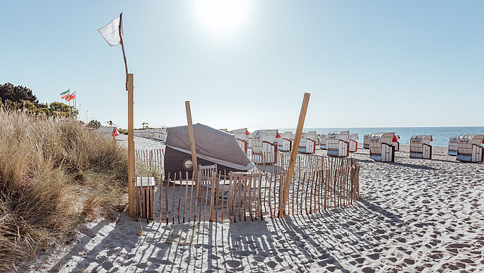 Der Schlafstrandkorb (geschlossen) steht auf dem Strand in der Nähe zum Deich und zur Ostsee ausgerichtet. Der Bereich um den Schlafstrandkorb ist mit einem filigranen Holzzaun von den restlichen Strandleben abgegrenzt.