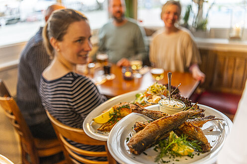 Vier Personen sitzen im Restaurant am Tisch. Der Kellner bringt das Essen. Auf dem Teller sieht man das Fischgericht.