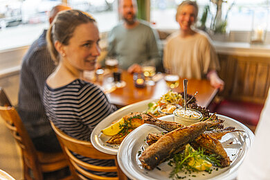 Vier Personen sitzen im Restaurant am Tisch. Der Kellner bringt das Essen. Auf dem Teller sieht man das Fischgericht.