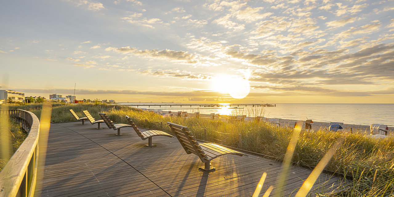 Landschaftsbild von der Promenade in Großenbrode bei Sonnenaufgang. Auf dem Holzdeck stehen drehbare Holzliegestühle