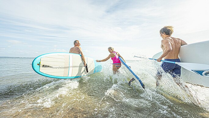 Eine Gruppe von Menschen mit SUP Boards unter dem Arm rennen ins Meer