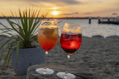 Zwei Cocktails stehen auf dem Baumstumpf, der als Tisch dient, im Sand. Zu sehen ist die Ostsee im Sonnenuntergang.