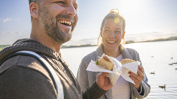 Eine Frau und ein Mann halten jeweils ein Fischbrötchen in der Hand und lächeln in die Kamera.