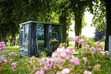 Im Vordergrund eine rosa blühende Blume. Ringsherum Kastanienbäume. In der Mitte eine Installation die ein großér Kasten ist. Dieser ist bemalt wie die Umgebung.
