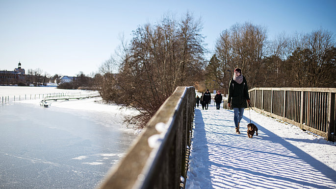 Menschen (warm angezogen) über die eingeschneite Brücke am Eutiner See. Der See ist an einigen Stellen gefroren und eine dünne Schneeschlicht liegt darüber.