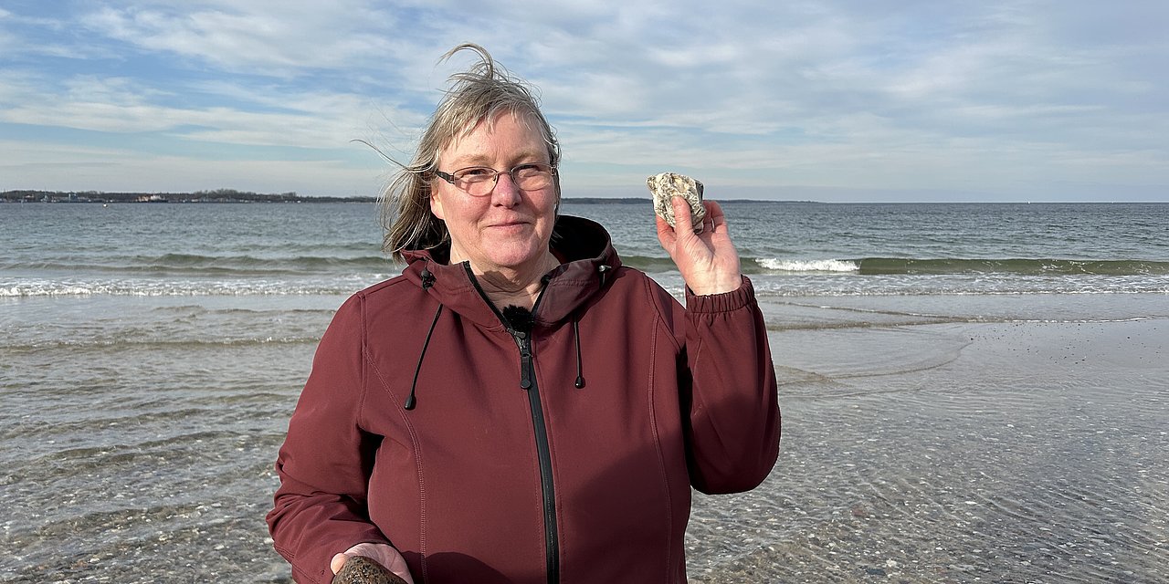 Eine Frau am Strand von Eckernförde. Sie hält einen Stein in die Kamera und lächelt. 