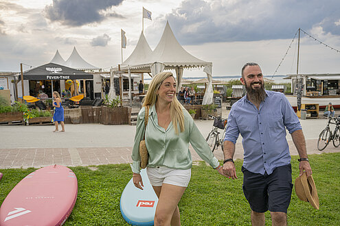 Ein Mann und eine Frau gehen Hand in Hand über ein Stück grüne Wiese von der Strandbar zum Strand. 
