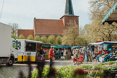 Menschen besuchen den Wochenmarkt in Neustadt. Auf dem Marktplatz stehen viele Verkaufswagen.