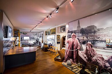 Ausstelungsstücke im Seebadmuseum. Dunkler Holzboden, an den Wänden sind Bilder und es sind Figuren aufgestellt die wie Seemänner aussehen.  