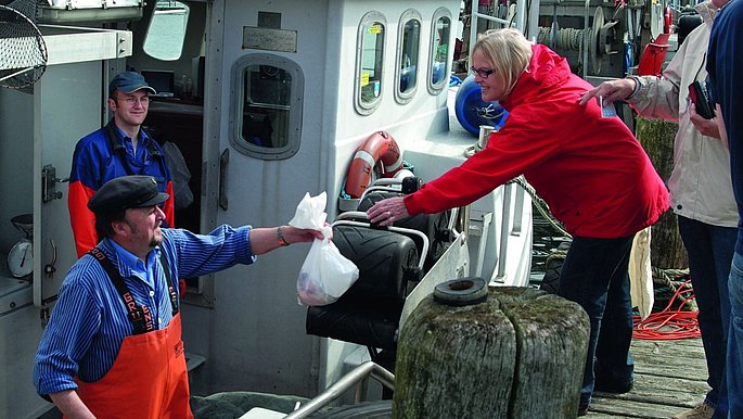 Ein Fischer verkauft Fisch an zwei Personen die vor seinem Boot stehen.