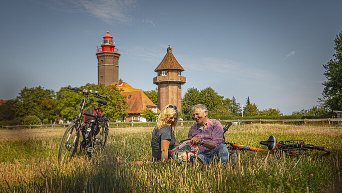 Vorne im Gras sitzt ein Pärchen. Links von ihnen stehen die Fahrräder. Im Hintergrund sieht man den Leuchtturm Dahmeshöved