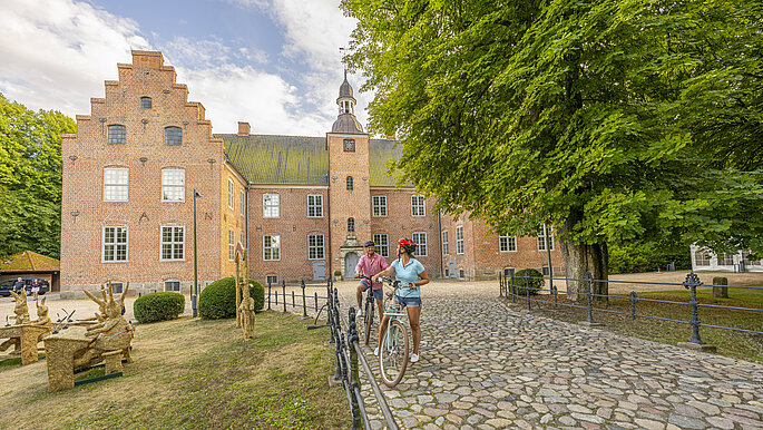 Ein Schloss im Hintergrund mit Backsteinen davor ein Weg über den zwei Menschen ihre Räder schieben. 
