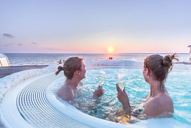 Zwei Freundinnen sitzen mit einem Glas Sekt im Whirlpool. Der Whirlpool steht in der Außenanlage der Ostsee Therme. Sie schauen beide in Richtung aufgehender Sonne am Horizont.