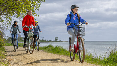 Radfahrer fahren auf einem Radweg direkt am Meer entlang. Im Vordergrund eine Frau mit Helm auf rotem Rad. Rechts im Bild das Meer.