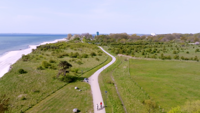 Ein Radweg von oben. Links sieht man das Meer, rechts grüne Wiesen. 