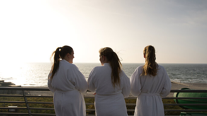 Drei Frauen stehen in weißen Bademänteln auf der Dachterrasse der Saunalandschaft. Sie schauen auf die Ostsee, die vor ihnen liegt. Der Betrachter des Bildes sieht die Frauen nur von hinten und blickt damit auch in die gleiche Richtung - die Ostsee.