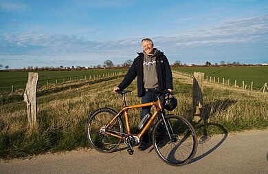 Axel Kramer steht mit seinem Fahrrad auf der Landstraße. Hinter ihm sieht man die Felder.
