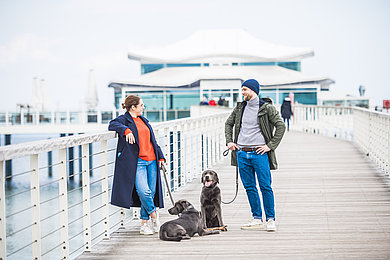 Zwei Personen stehen auf der teehaus-Seebrücke in Timmendorfer Strand. Davor sitzt ein Hund und einer liegt. 