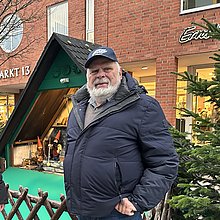 eine Frau und ein alter Mann vor einer hölzenern Hütte, in der Zwerge aufgestellt sind, auf einem Weihnachtsmarkt. 