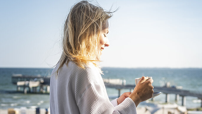 Frau steht im Bademantel auf dem Balkon und genießt ihren Kaffee mit Ausblick auf die Seebrücke Heiligenhafen und die Ostsee.